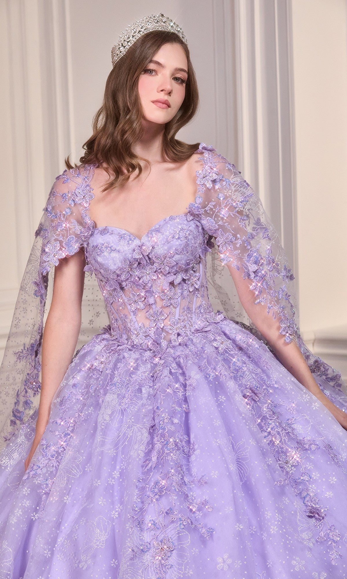 A woman in the Ladivine Quinceanera Dress 15719 in lavender, featuring floral appliques and a sheer cape, poses indoors wearing a jeweled crown.
